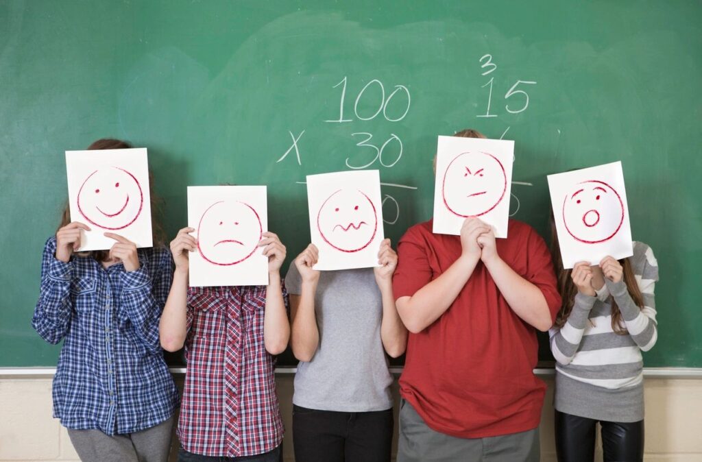 Five children in front of chalkboard holding up drawn faces in front of their own, depicting different emotions.